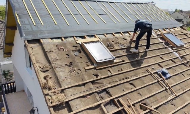 Un homme travaille sur le toit d'une maison, en retirant des planches et des fenêtres de toit.