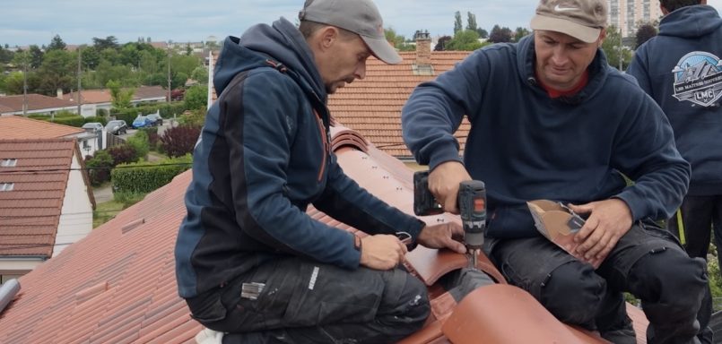 Deux ouvriers posent des tuiles sur un toit. Ils portent des casquettes et des vêtements de travail.