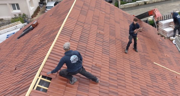 Deux travailleurs posent des tuiles sur un toit en pente. Outils visibles autour d'eux.